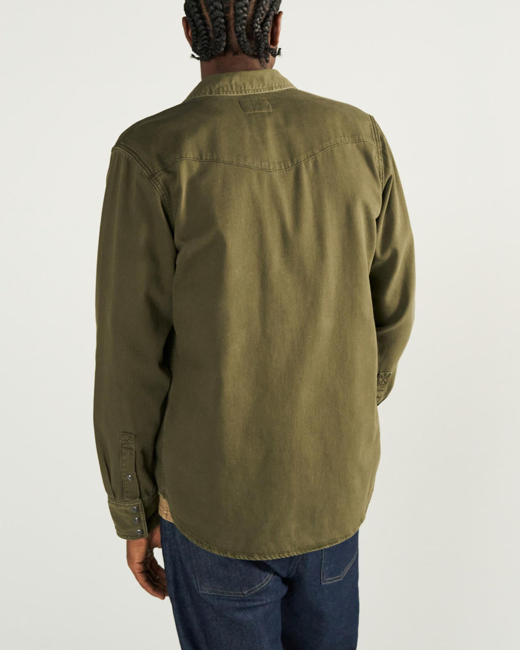 A person with braided hair is shown from behind, wearing an olive green long-sleeve shirt and dark blue jeans against a plain light background.