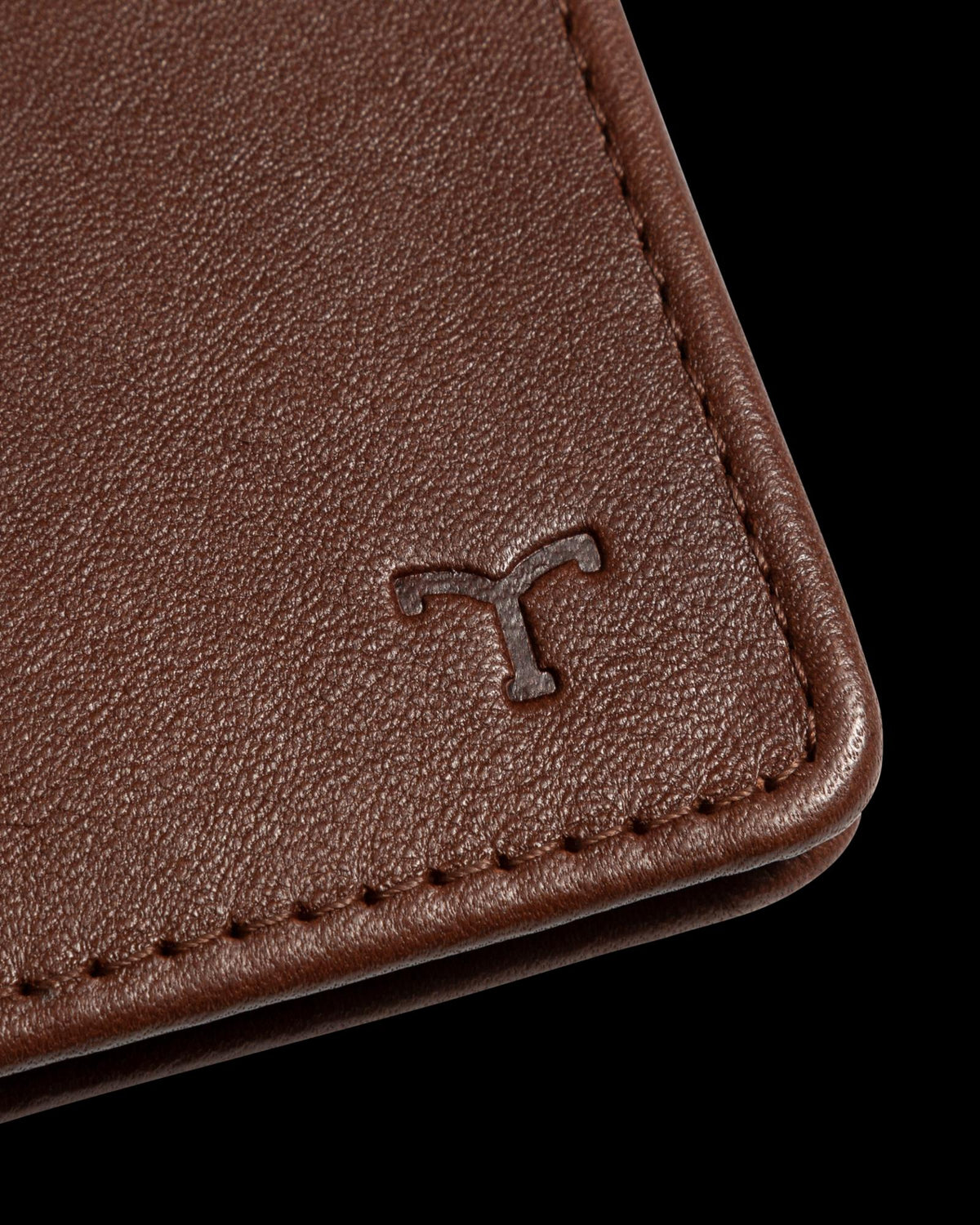 Close-up of a brown leather wallet corner featuring a small embossed logo resembling the letter "T.