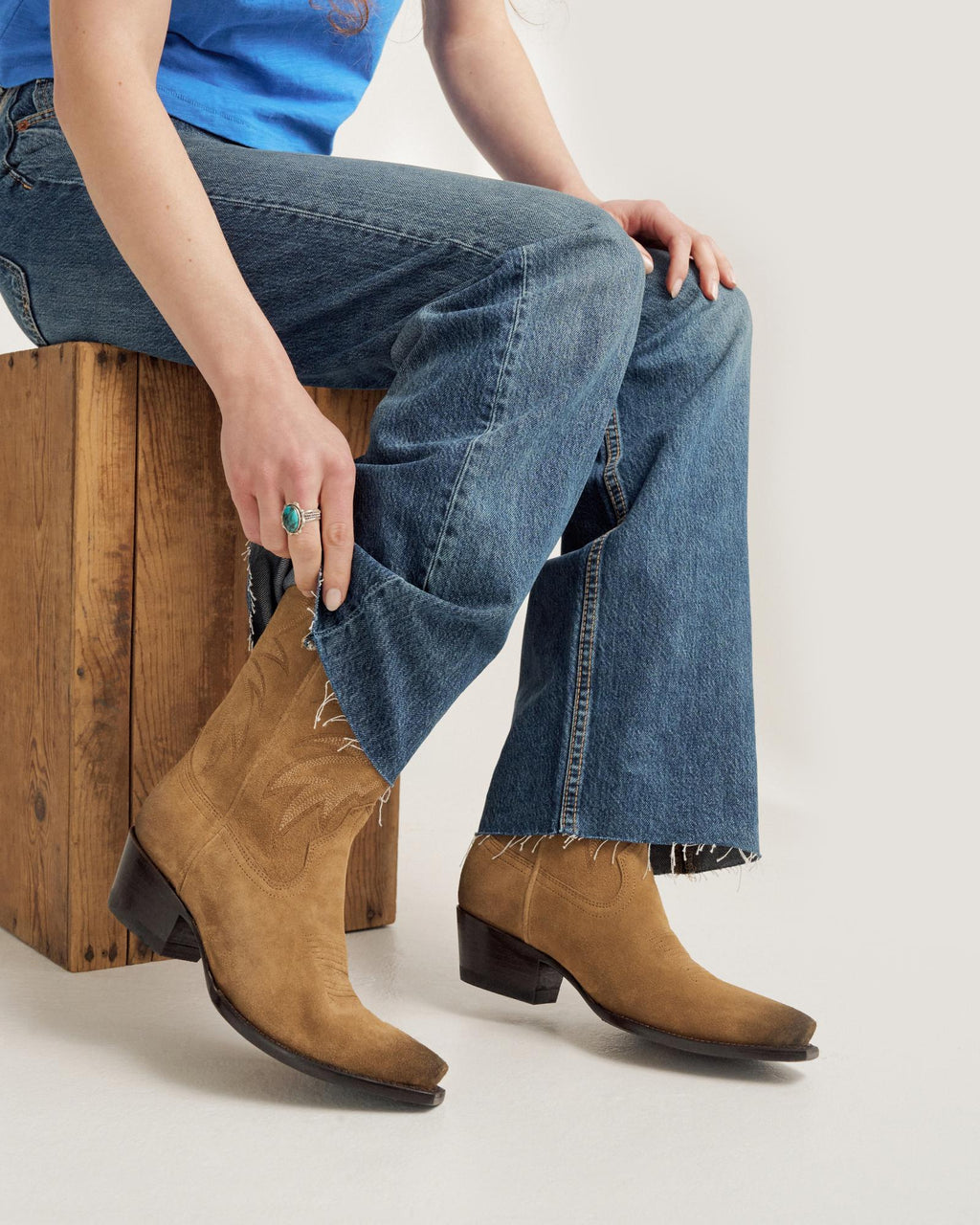 A person sitting on a wooden box wearing blue jeans, a blue shirt, and tan suede cowboy boots with a pointed toe and stacked heel.