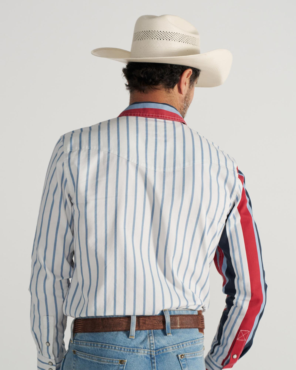 A man wearing a white cowboy hat, a striped long-sleeve shirt with red accents, blue jeans, and a brown belt stands facing away from the camera.