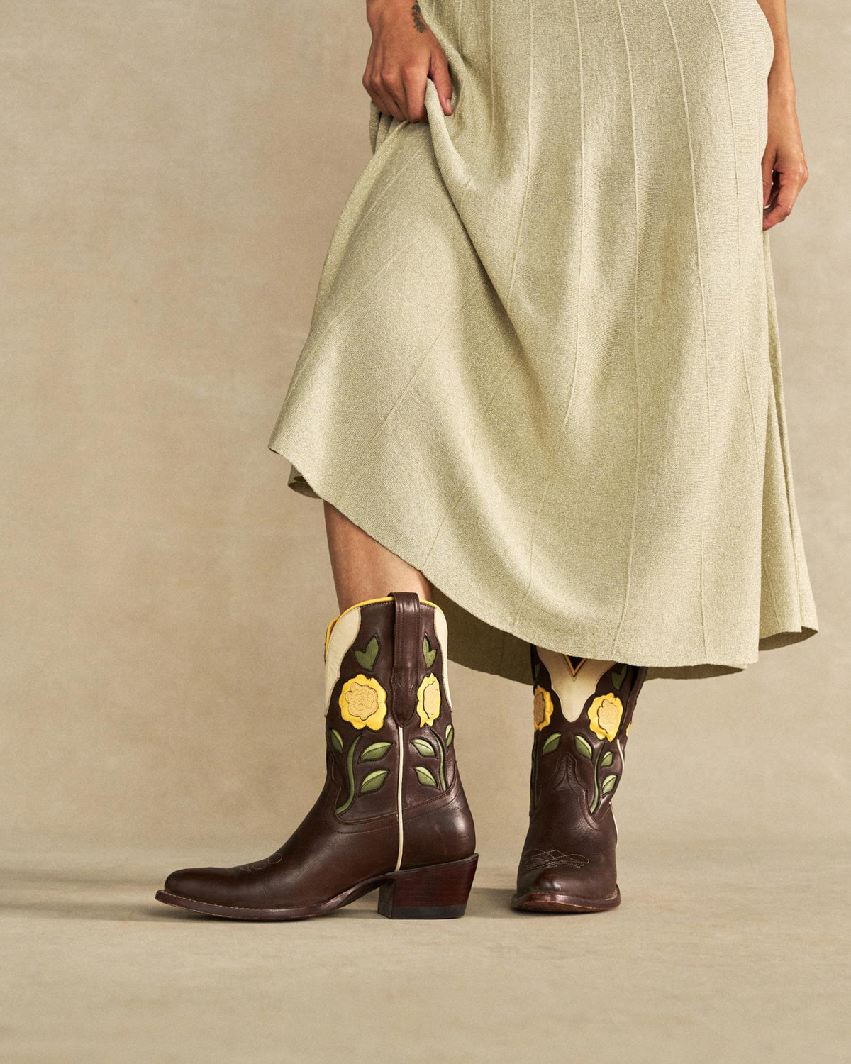 The Jolene in Truffle Cowhide. A person in a light-colored skirt displays The Jolene Cowhide brown cowboy boots decorated with yellow flower and green leaf designs, standing on a neutral backdrop.