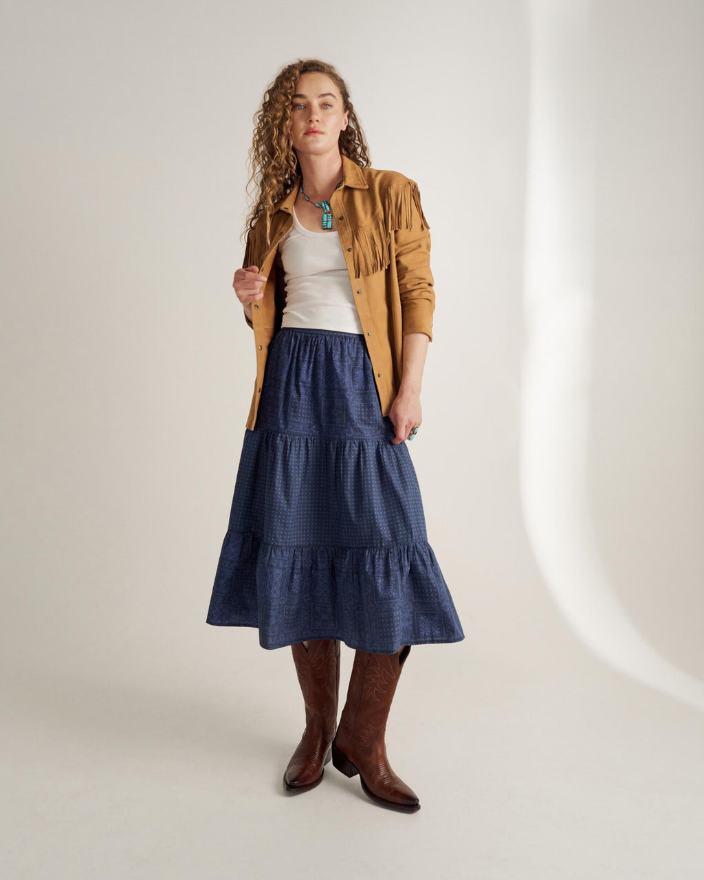 A woman stands in a studio wearing a tan fringe jacket, white top, tiered blue skirt, and brown cowboy boots.
