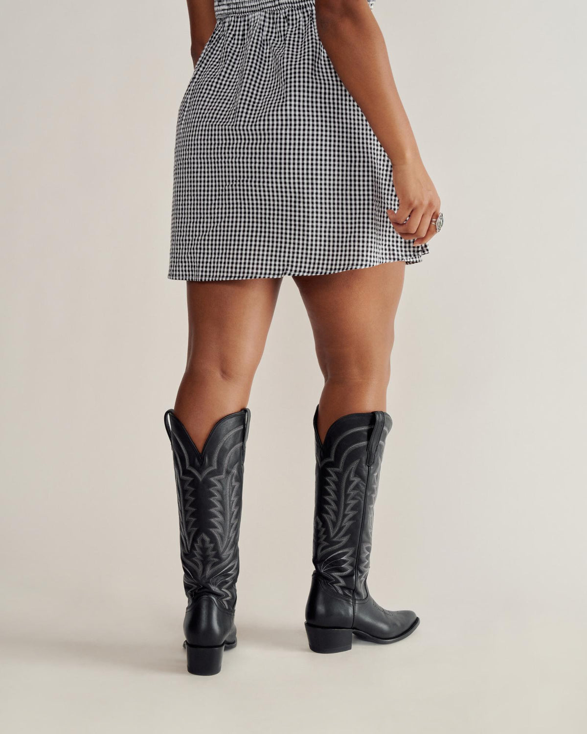 A person wearing a black and white checkered dress and black cowboy boots stands facing away, with one hand by their side.