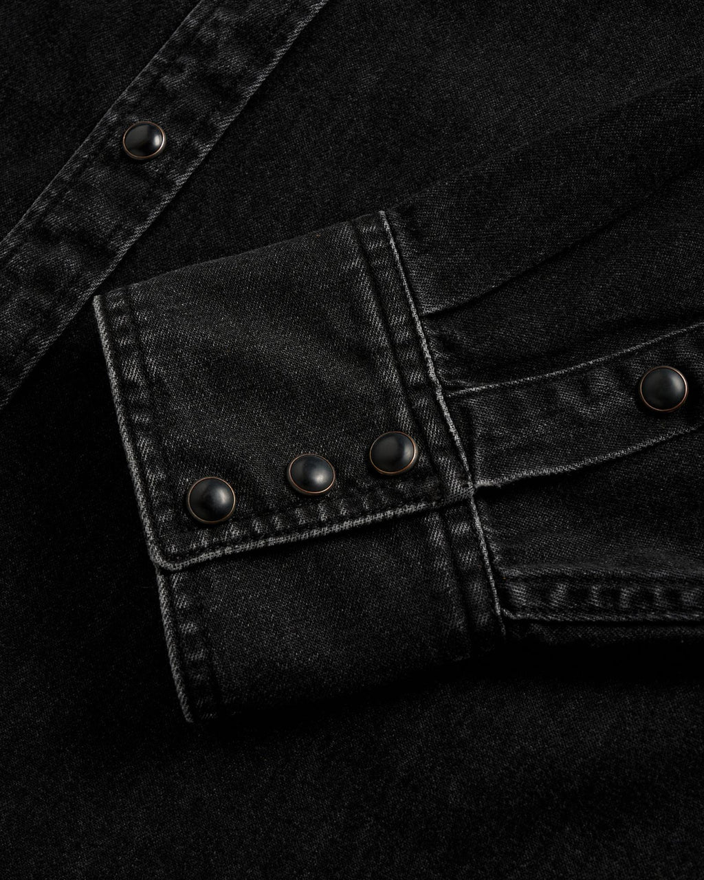 Close-up of a black denim shirt cuff with three black snap buttons and visible white stitching.