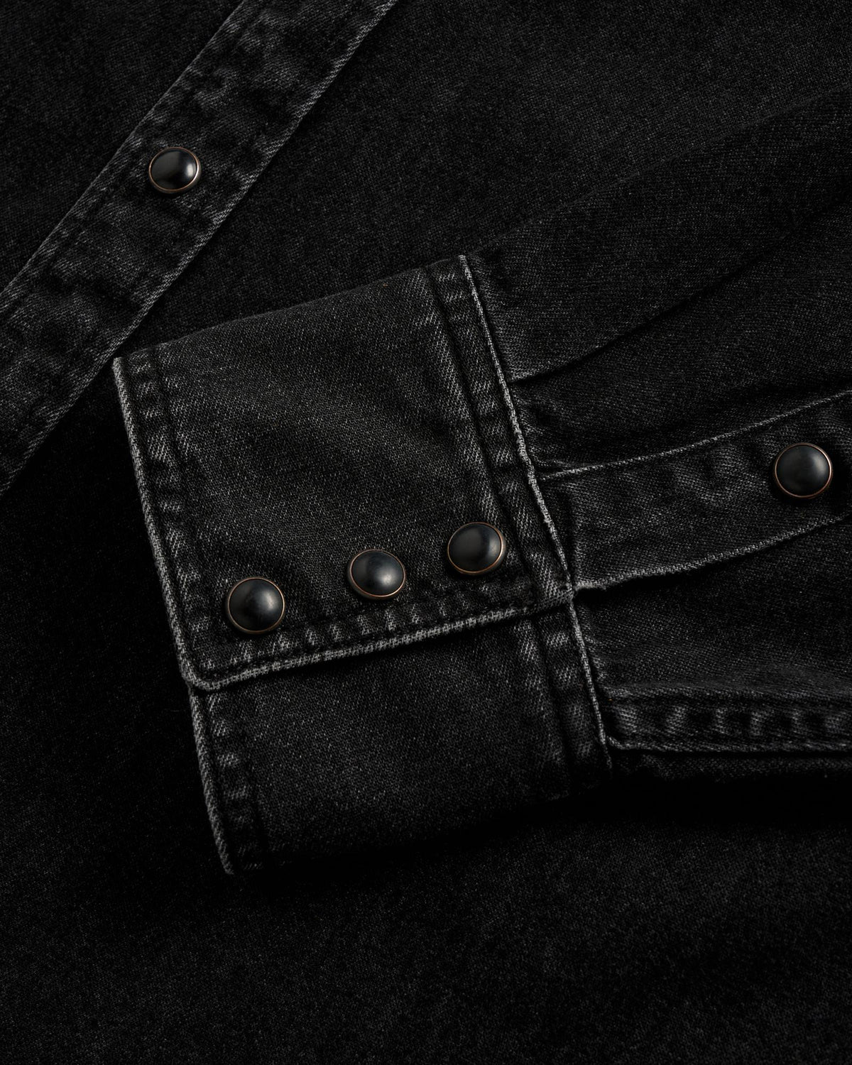 Close-up of a black denim shirt cuff with three black snap buttons and visible white stitching.