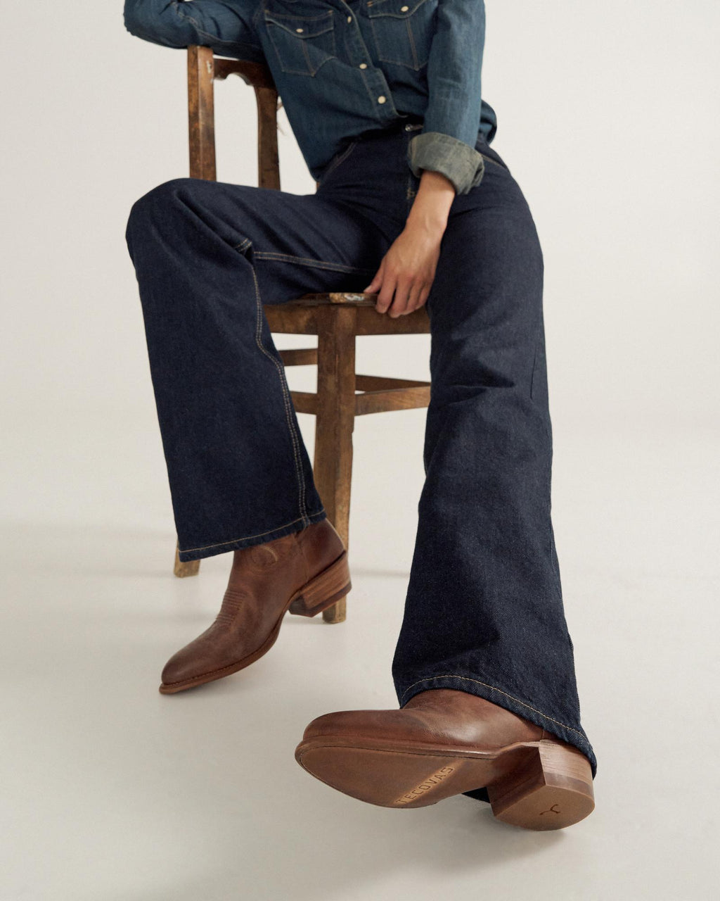 A person sits on a wooden chair wearing a denim shirt, dark flared jeans, and brown leather western boots, with one arm resting on the chair back.