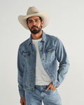 A man wearing a white cowboy hat, a light blue denim jacket over a white shirt, and blue jeans stands against a plain light background.