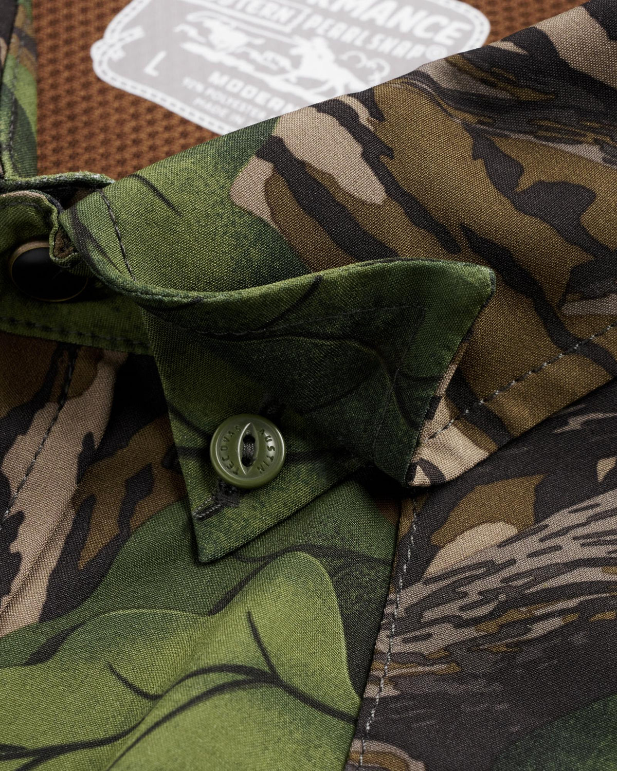 Close-up of a camouflage shirt collar with a green button, featuring mixed camo patterns in green, brown, and black, with a size label visible in the background.