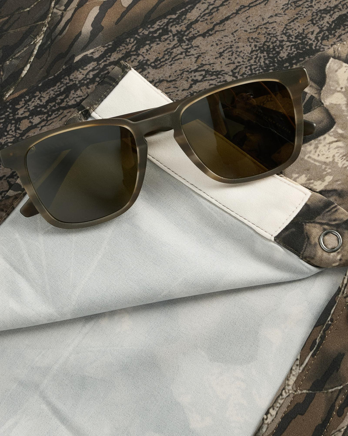 A pair of brown sunglasses rests on a folded white and camouflage-patterned cloth with a metal button.