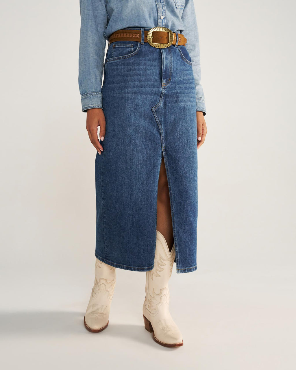 A person wearing a blue denim skirt with a front slit, a brown belt, a denim shirt, and white cowboy boots stands against a plain background.