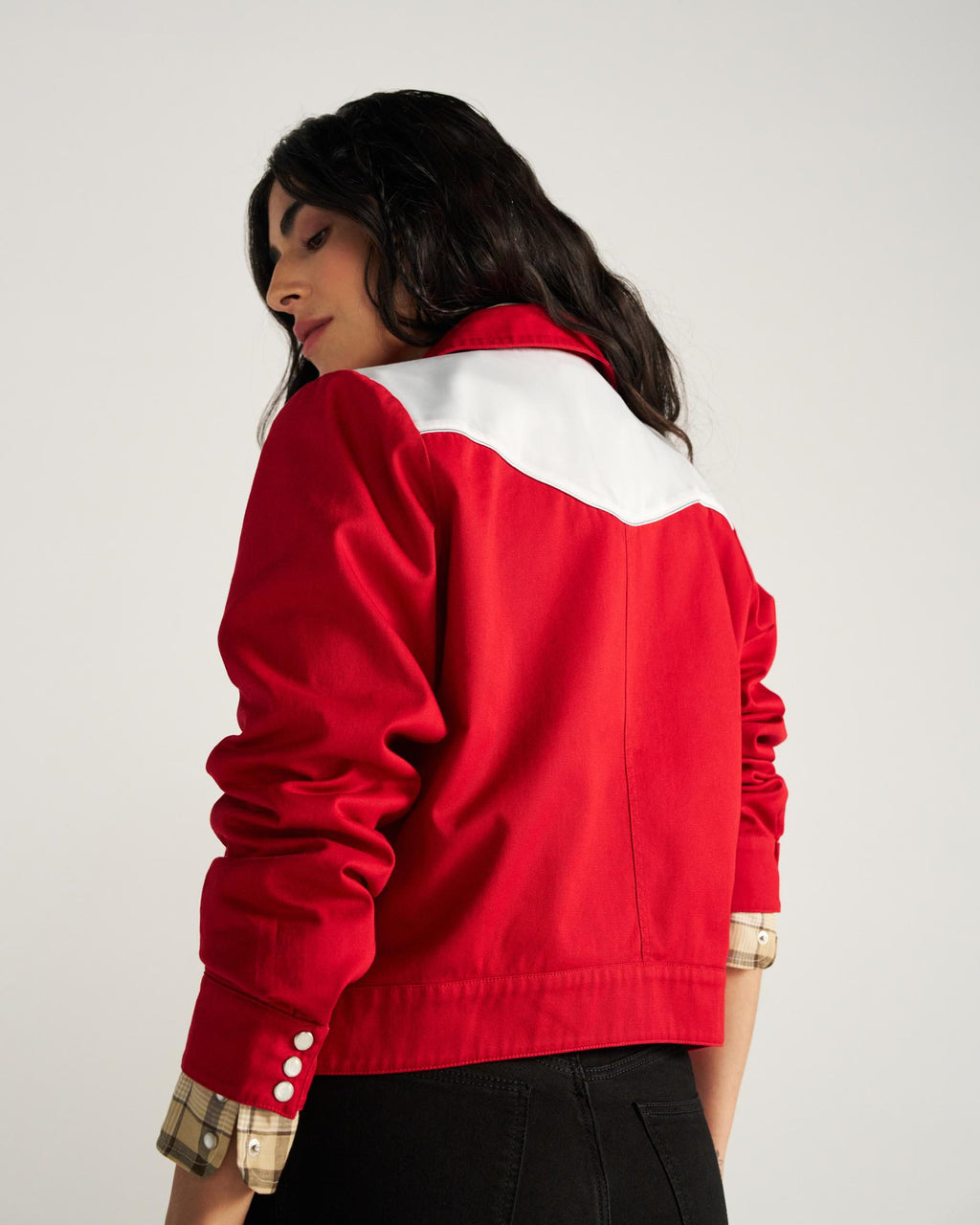 A person with long dark hair wears a red jacket with a white yoke, standing with their back to the camera against a plain background.