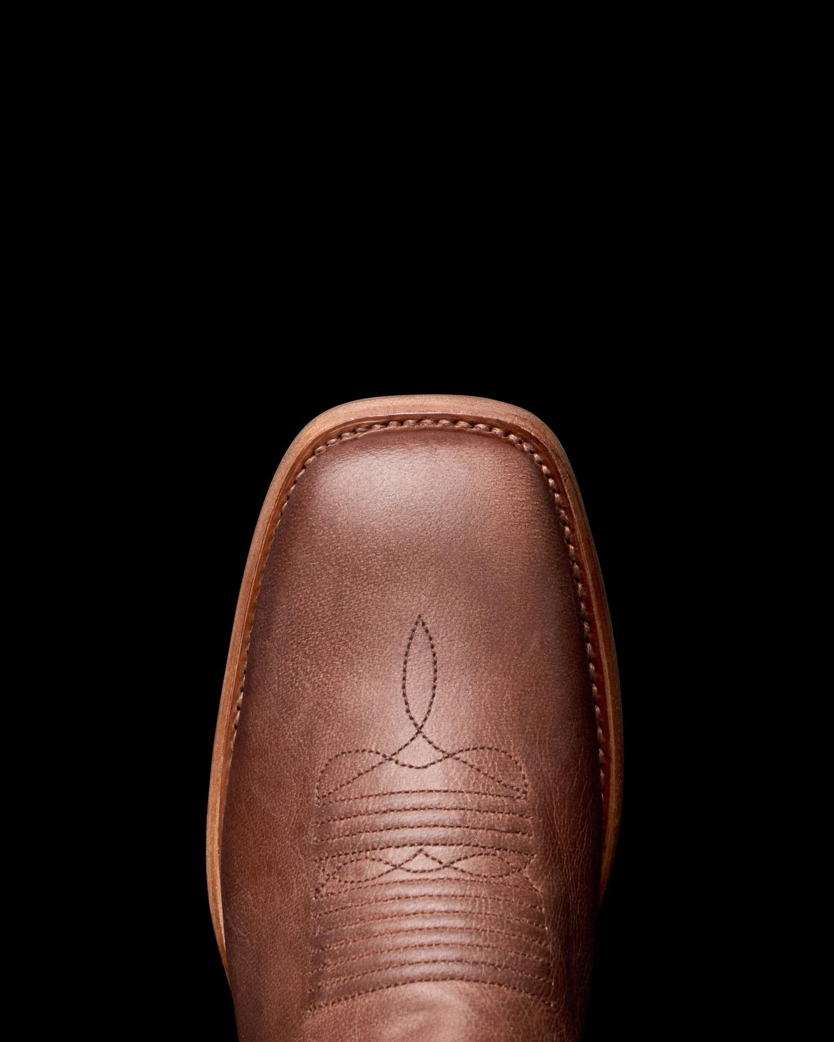 Close-up of a brown leather cowboy boot with decorative stitching on a black background.