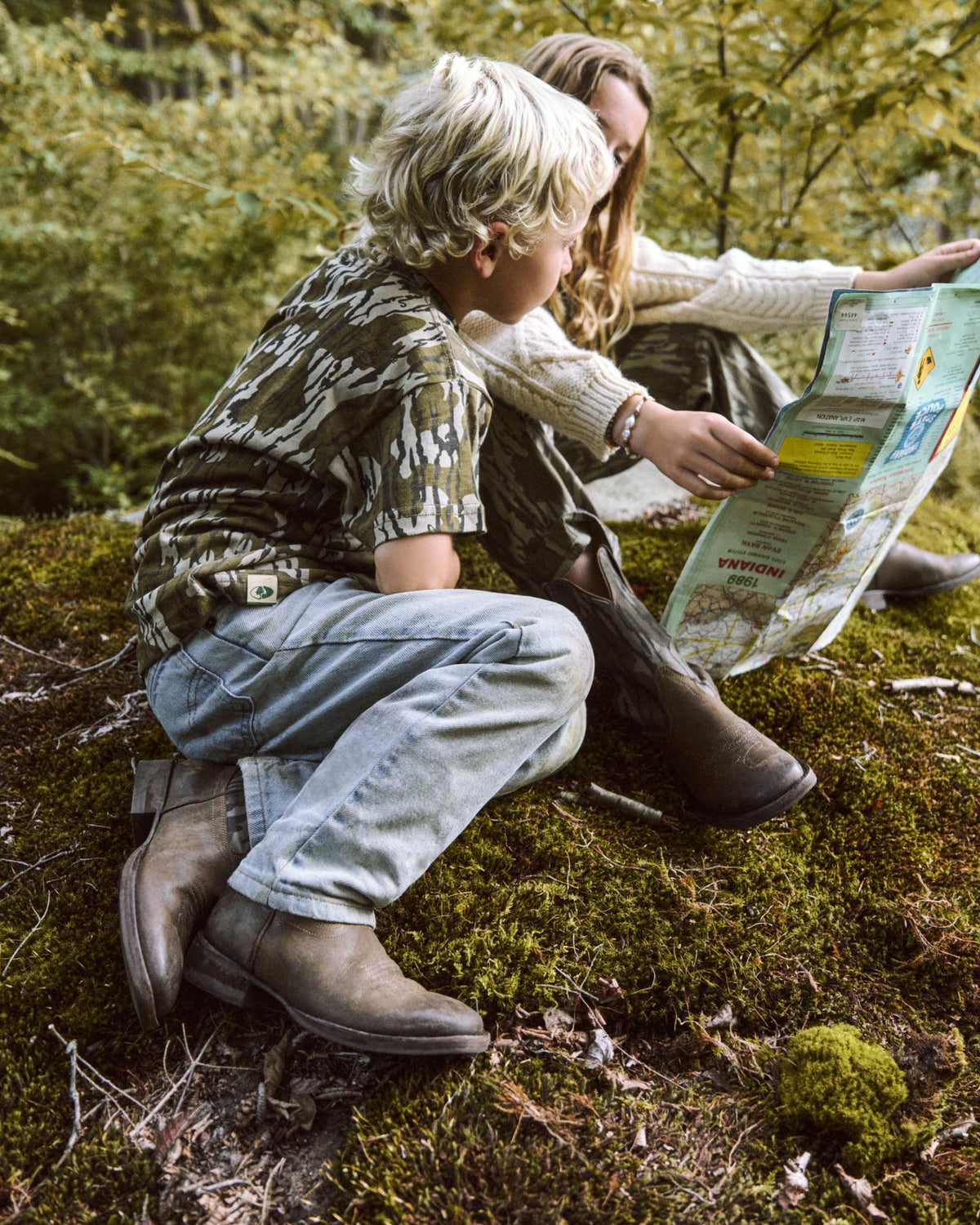 A child and an adult sit on mossy ground outdoors, looking at a map together; trees and greenery surround them.