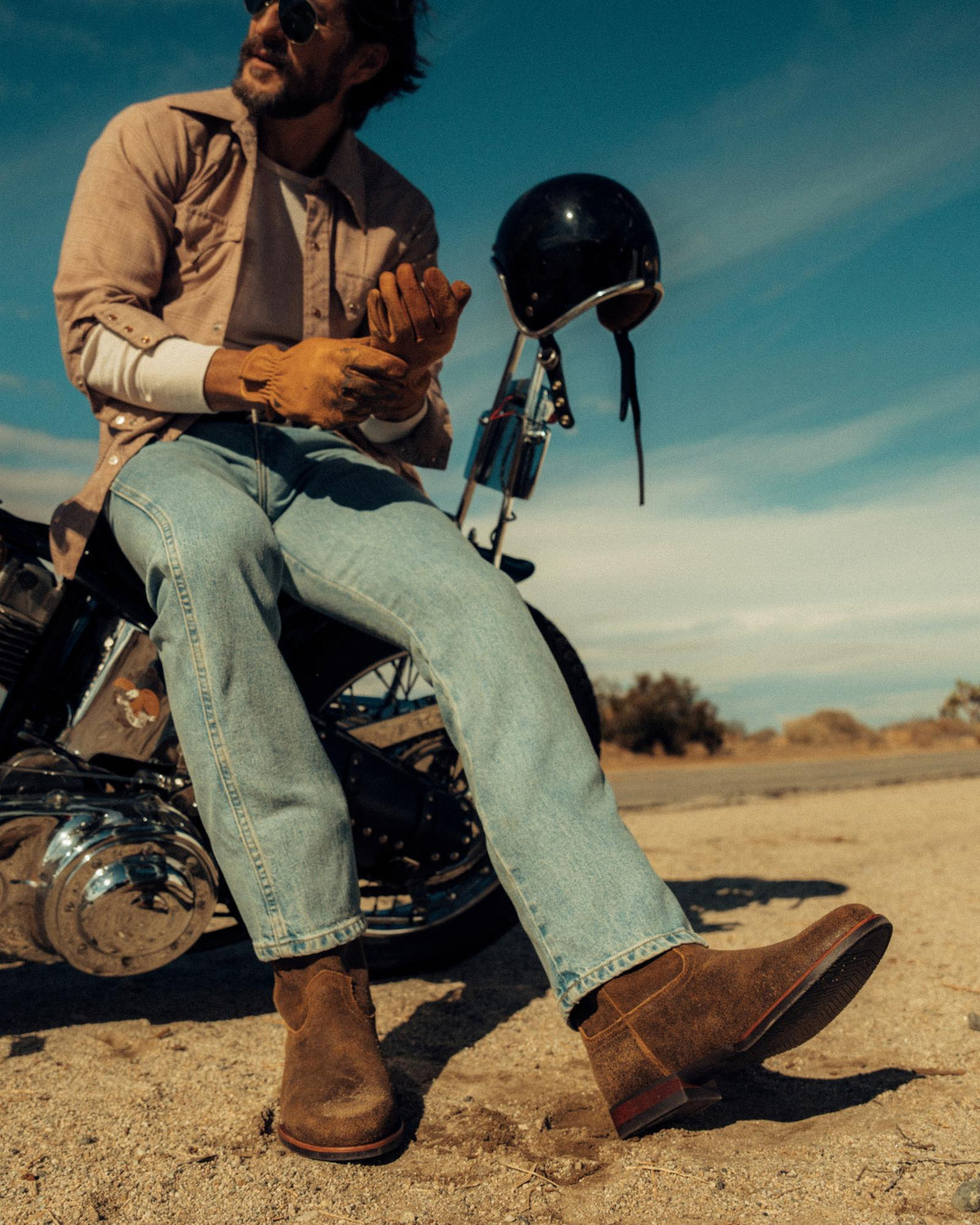 The Glen in Dustwood Roughout. Man sitting on a Dustwood Roughout motorcycle, wearing a light shirt, jeans, gloves, and boots, with a helmet hanging on the bike under a clear sky in a desert setting.