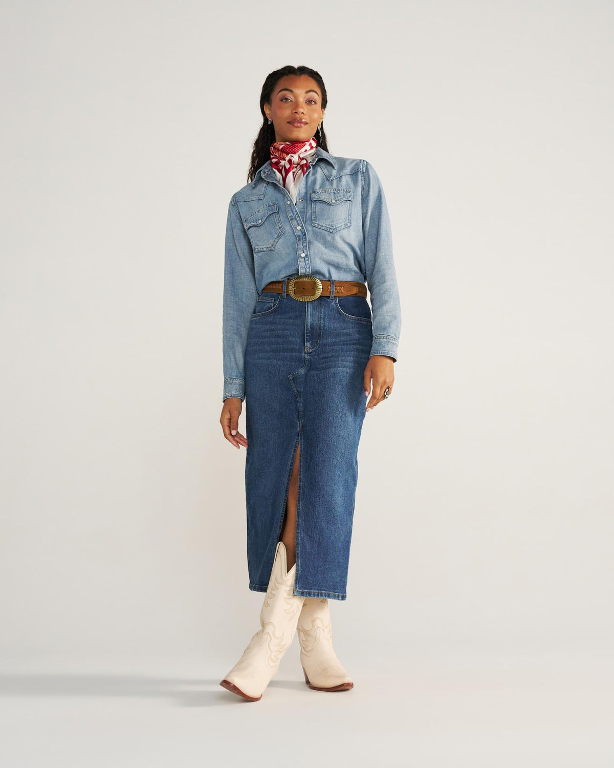 A woman stands wearing a denim shirt, denim skirt with a front slit, white cowboy boots, a brown belt, and a red patterned neck scarf.