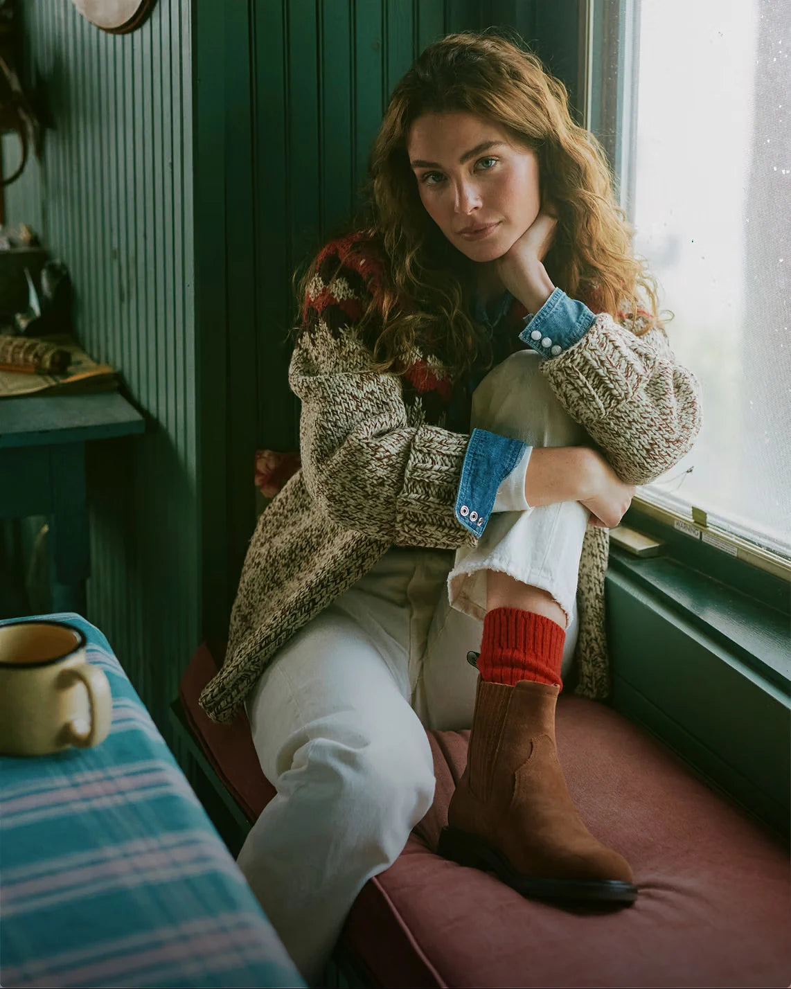 Layered knitwear and denim by a sunlit window